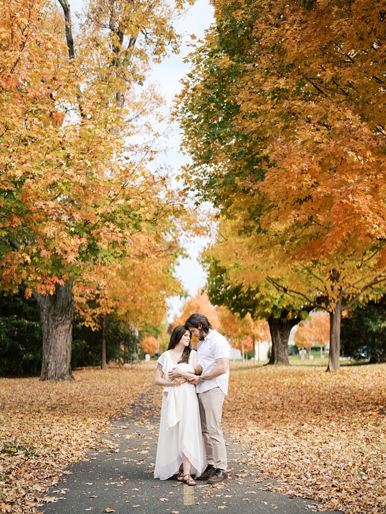 A Young Couple Stand Among A Row Of Orange Maple Trees During Their Newborn Session In Rockville, Maryland.