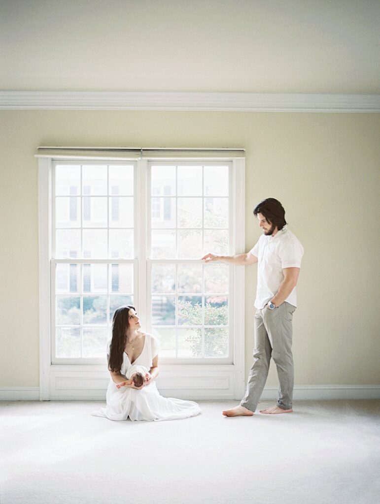 A Mother Sits In Front Of The Window Holding A Newborn Baby As A Father Stands Nearby Looking On.