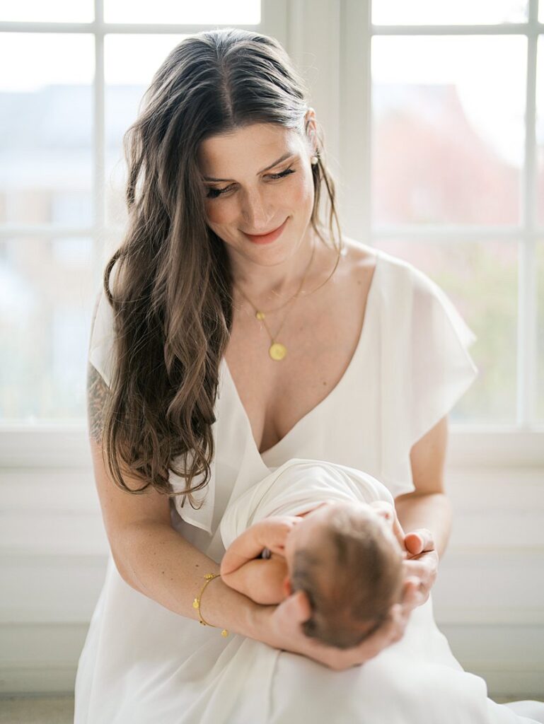 A Mother With Long Brown Hair Holds Her New Baby In Front Of Her As She Sits In Front Of The Window.