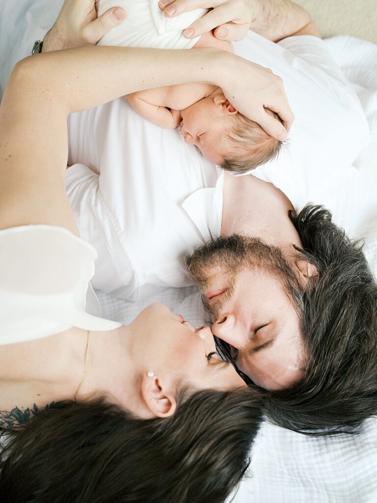 Brown-Haired Mother And Father Lay Down Nose-To-Nose With Their Baby On The Father's Chest.