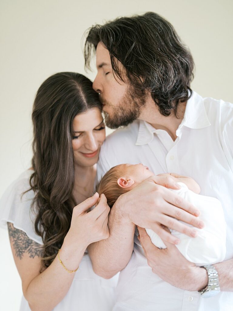 A Father Kisses His Wife's Head As He Holds Their Newborn Baby During Their Rockville Newborn Photography Session.