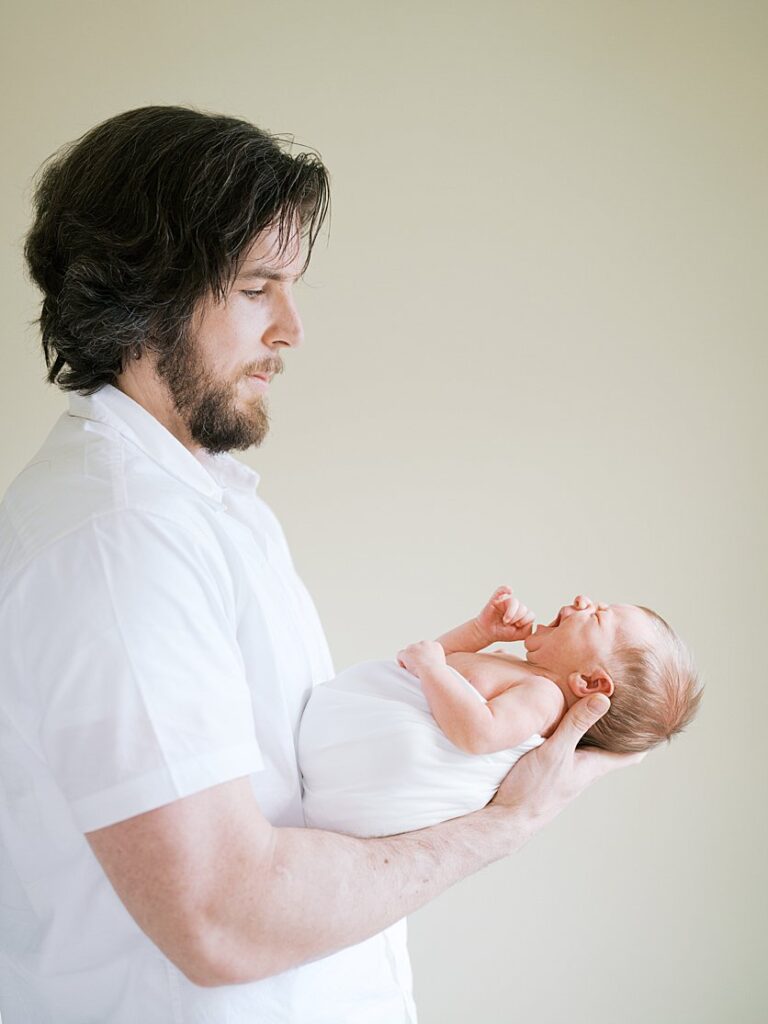 Father With Brown Hair And A Beard Holds His Newborn Baby In Front Of Him.