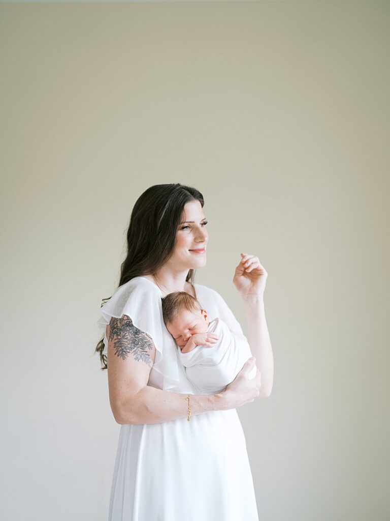 Mother With Brown Hair Stands With Her Newborn Against Her Chest And A Soft Smile On Her Face.