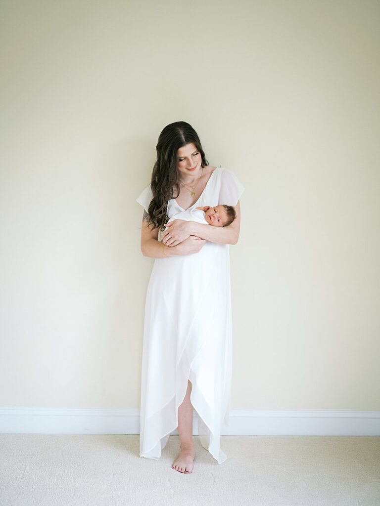 A Mother With Long Brown Hair Leans Against A Wall While Holding Her Newborn Son.