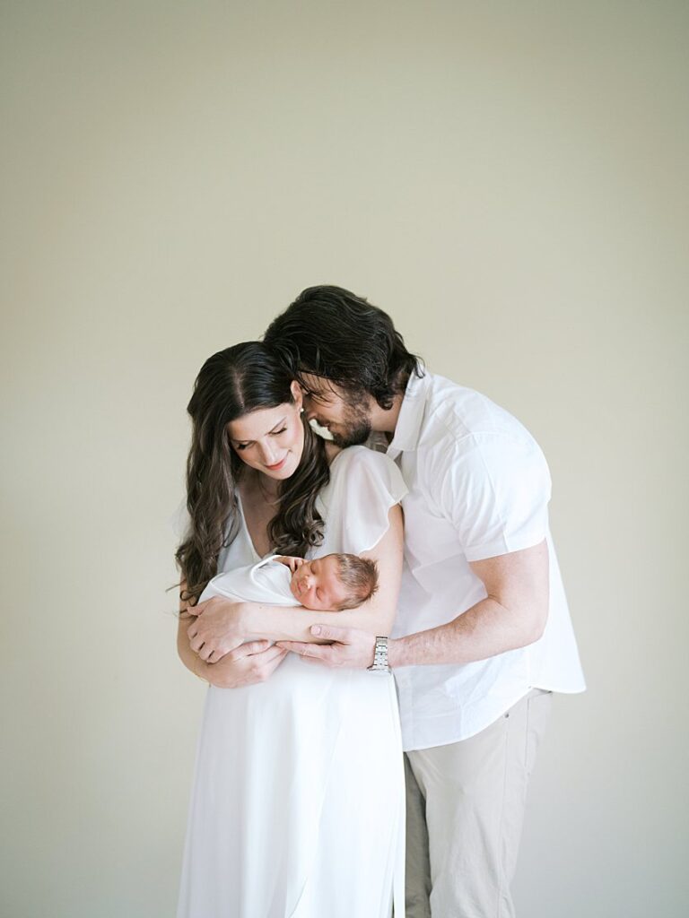 A New Father Leans Into His Wife As She Smiles Down At Their Newborn Baby During Their Rockville Newborn Photography Session.