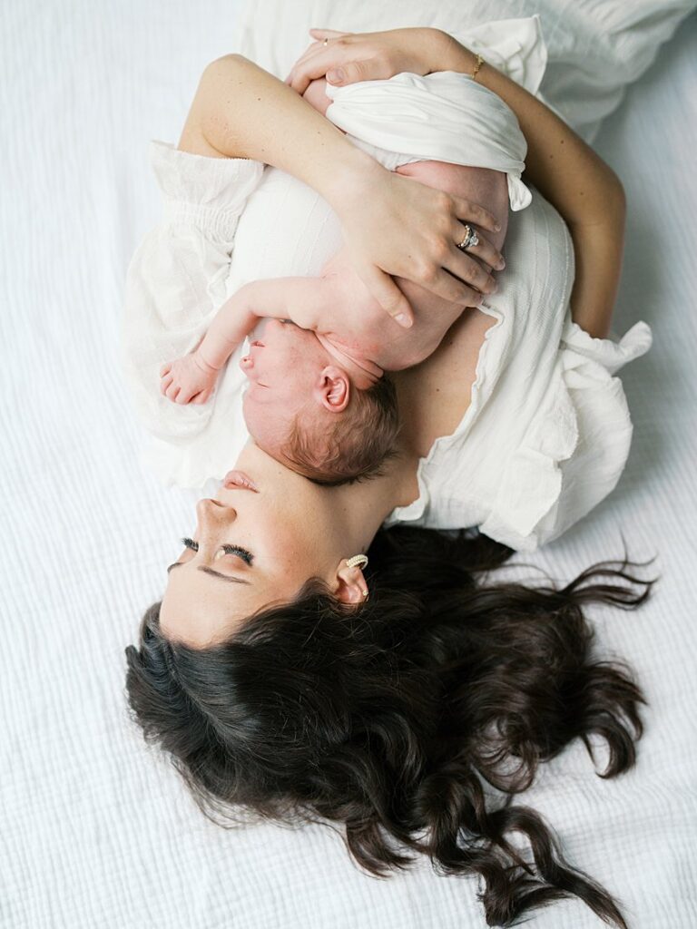 A Mother With Brown Hair Lays On The Ground Holding Her Newborn Baby On Her Chest.