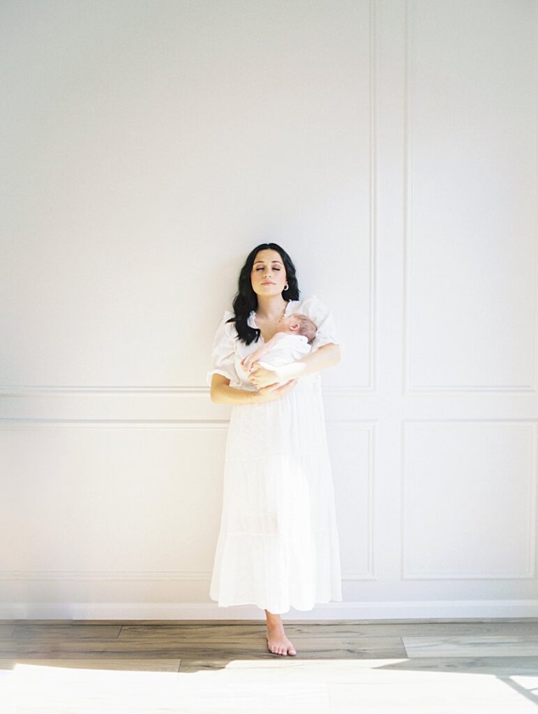 A Mother With Long Brown Hair Leans Against A White Wall In Her Dining Room Holding Her Newborn Baby During Their Severna Park Newborn Photos.