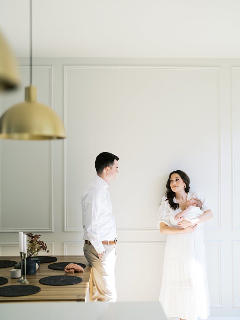 A Brown-Haired Mother Leans Up Against Her White Wall In Her Dining Room Holding Her Newborn Looking At Her Husband During Their Severna Park Newborn Photos.