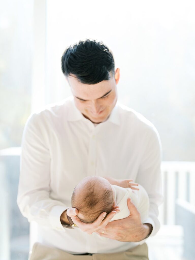 Father With Brown Hair Looks Down And Smiles At His Newborn Baby.