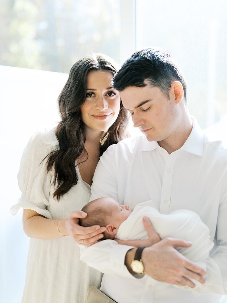 Mother With Brown Hair And Eyes Looks At The Camera As Her Husband Holds Their Newborn Baby.