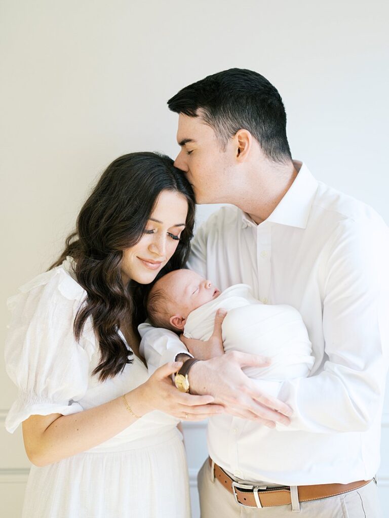 A Father Holds His Newborn Son And Kisses His Wife's Head During Their Severna Park Newborn Photos.