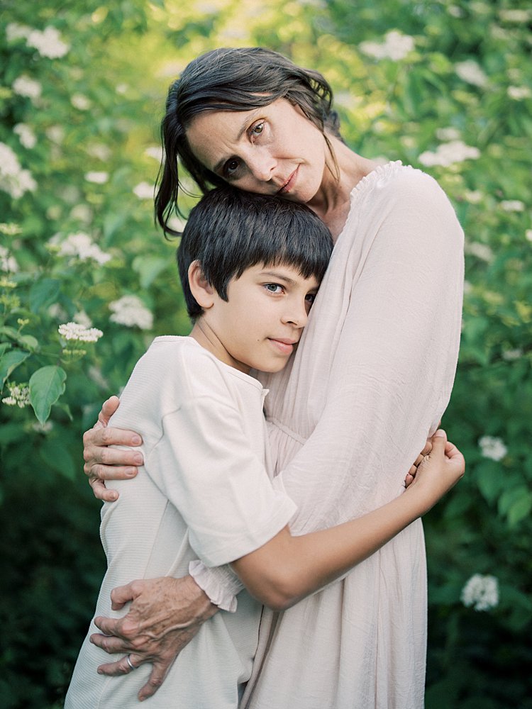 A Mother With Brown Hair Embraces Her Son As They Stand In Front Of White Flowers During Their Great Falls Family Photos.