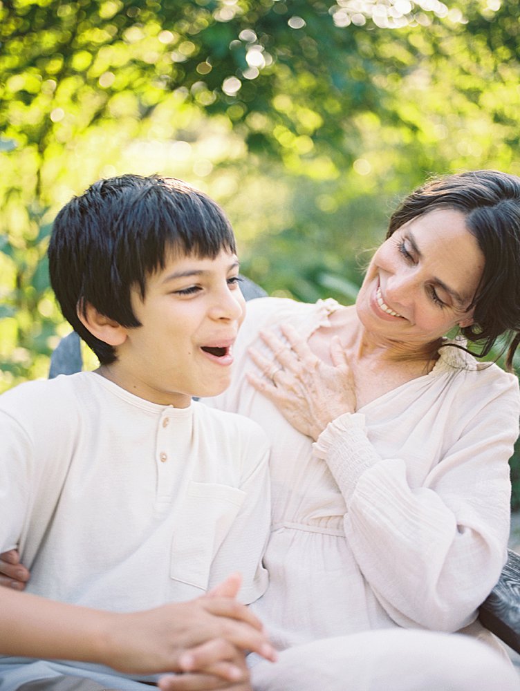 A Boy And His Mother Sit In Front Of Greenery And Laugh.