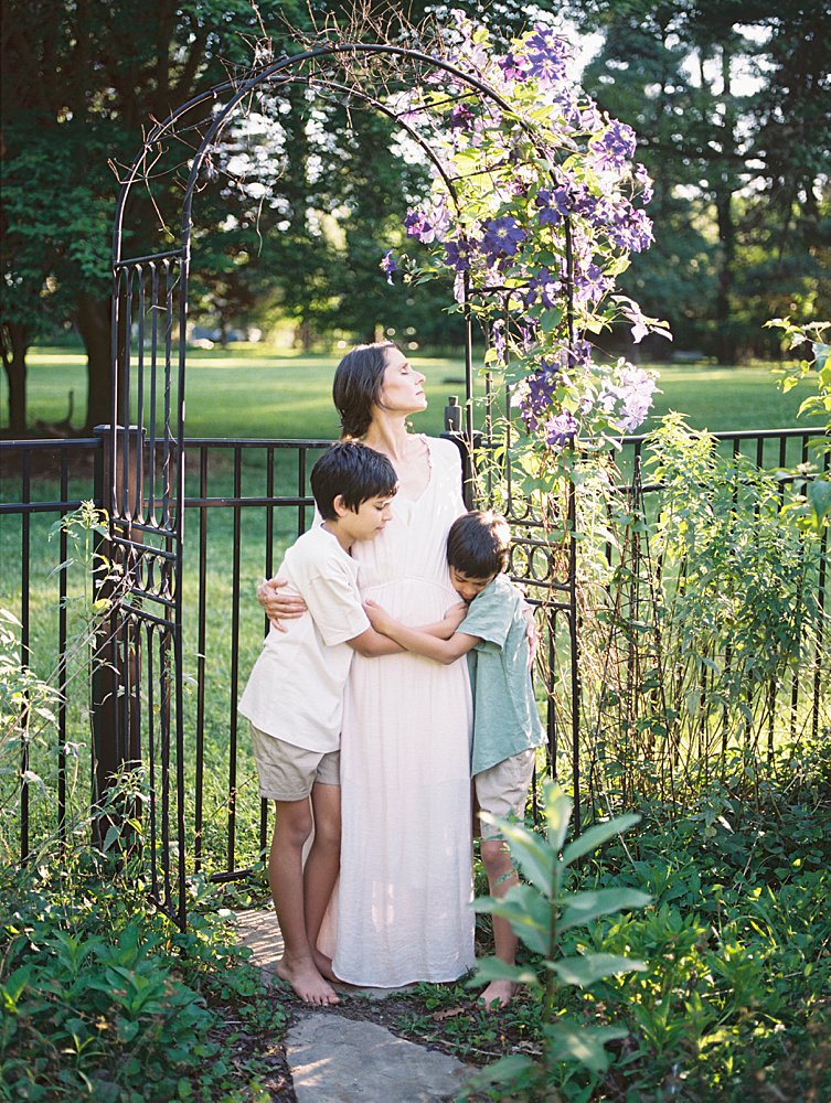 A Mother In A Long Ivory Dress Stands Under A Purple Floral Arch While Embracing Her Sons During Their Great Falls Family Photos.