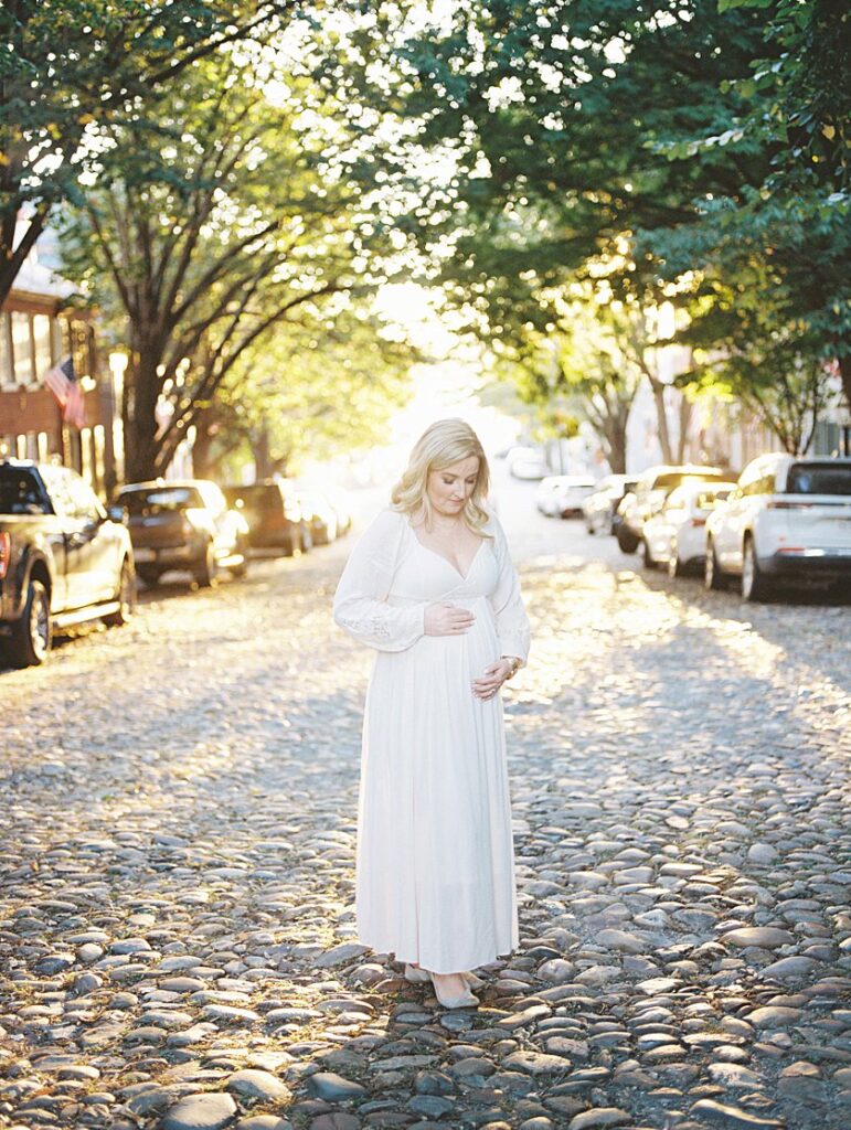 A Blonde Mother-To-Be Stands With Her Hands On Her Belly On The Cobblestone Street Of Prince Street - Maternity Photography In Alexandria Va By Marie Elizabeth Photography.