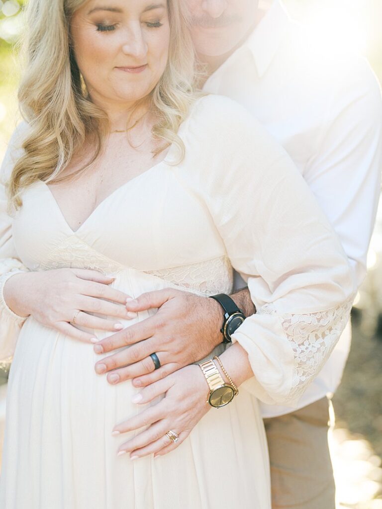 Close-Up View Of An Expecting Couple Standing Together Against The Sunlight With Hands On The Mom's Belly.