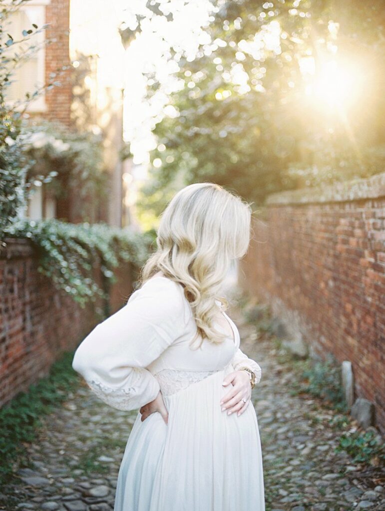 A Pregnant Mother Stands In An Alley With A Hand On Her Belly And One Hand On Her Back During Sunrise - Maternity Photography In Alexandria Va By Marie Elizabeth Photography.