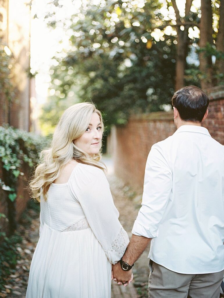 A Blonde-Haired Woman Turns To Look Back At The Camera As She Walks Down An Alley In Old Town Alexandria With Her Husband.