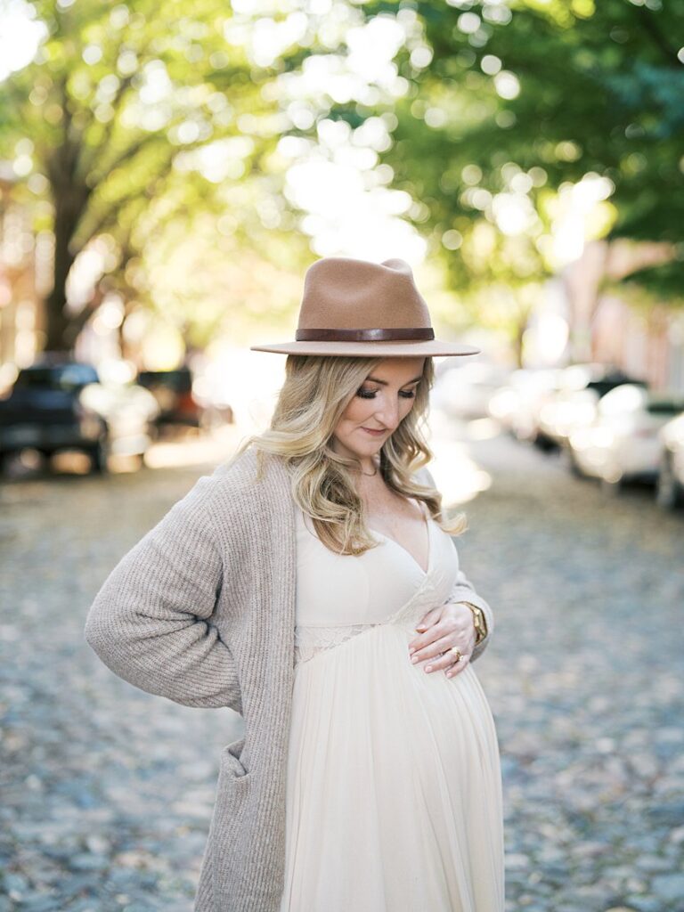 Mother In An Ivory Dress, Taupe Sweater And Hat Stands On Prince Street With A Hand On Her Belly - Maternity Photography In Alexandria Va By Marie Elizabeth Photography.