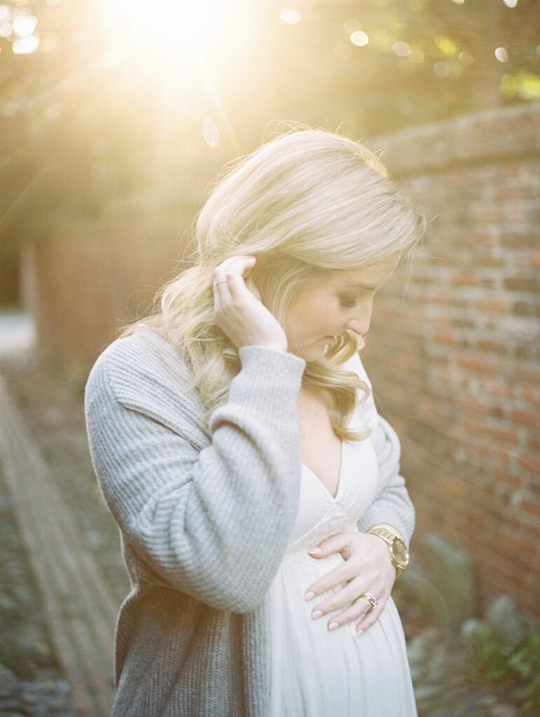 A Mother Brushes Away Hair By Her Ear As She Places A Hand On Her Pregnant Belly During Her Maternity Session.