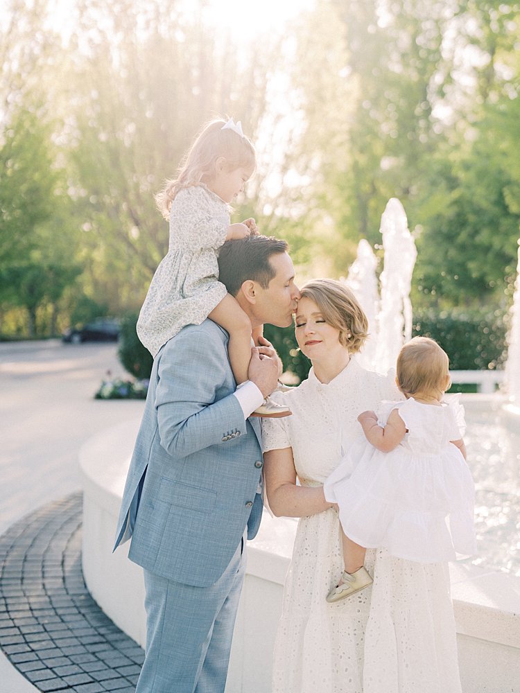 A Little Girl Sits On Her Father's Shoulders As He Leans Over To Kiss His Wife's Head During Their Mormon Temple Washington Dc Family Photos.