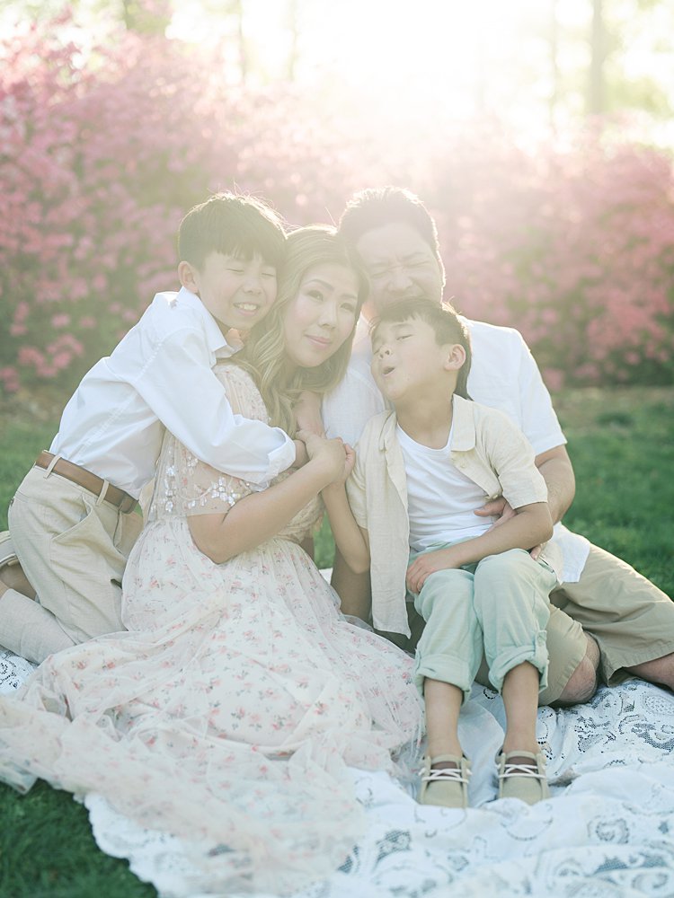 A Family Of Four Sits On A Blanket Close Together While The Mother Looks At The Camera During Sunset At Brighton Dam Azalea Garden.