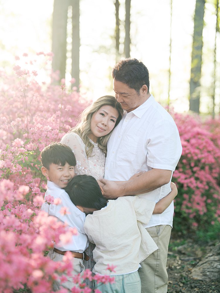 Mother Looks At The Camera As She Leans Into Her Husband And Her Two Sons Hug Her In Front Of Pink Azaleas.