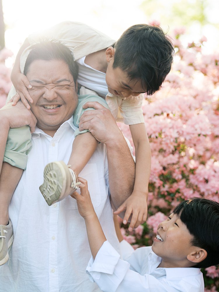 One Boy Sits On His Father's Shoulders As He Leans Down To Tickle His Younger Brother.