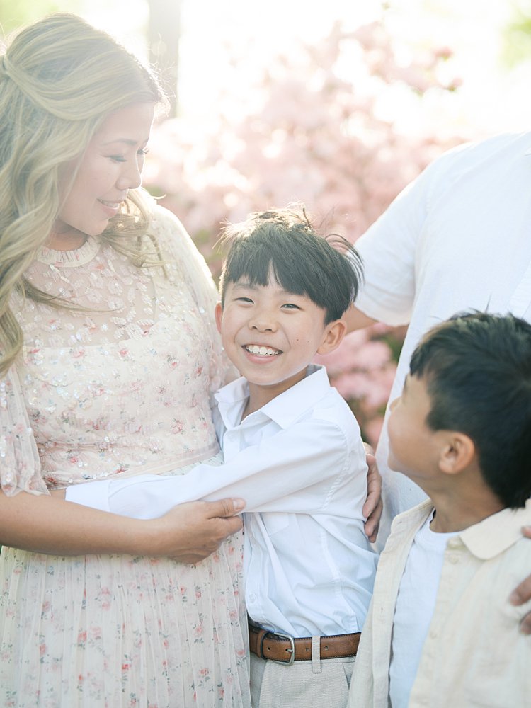 Closeup View Of A Little Boy Smiling At The Camera While His Mother And Brother Look Adoringly At Him.