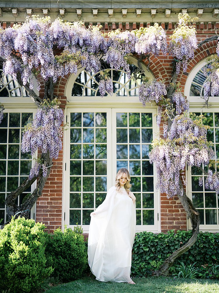 A Blonde Pregnant Mother, Sways Her White Dress Against A Brick Building With Wisteria At Dumbarton Oaks, Photographed By Georgetown Photographer Marie Elizabeth Photography.