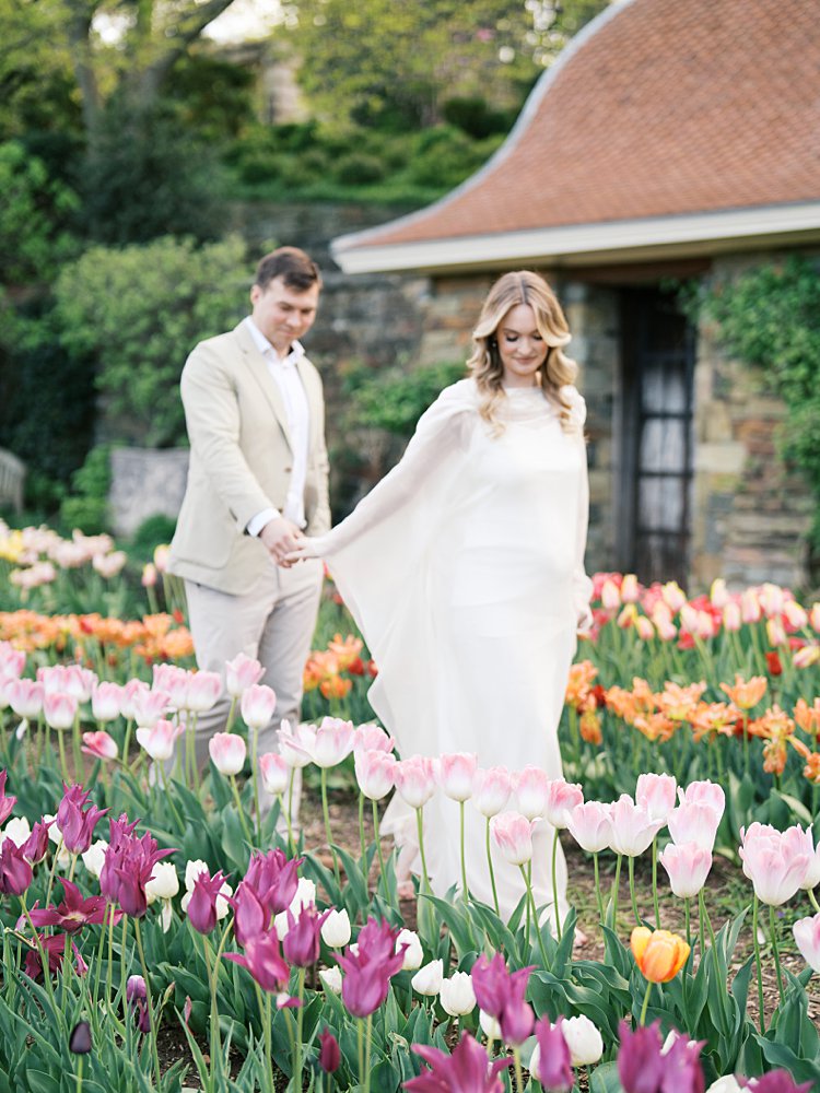 A Couple Walk Through A Field Of Tulips Together During Their Maternity Session At Dumbarton Oaks Garden, Photographed By Georgetown Photographer Marie Elizabeth Photography.