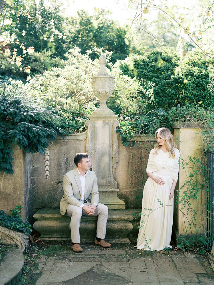 A Pregnant Mother Leans Against A Wall, Looking At Her Husband As He Sits On A Stone Bench In Dumbarton Oaks.