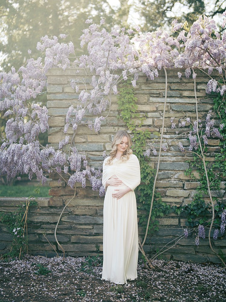 A Pregnant Woman Stands Along A Stone Wall Covered In Wisteria, Photographed By Georgetown Photographer Marie Elizabeth Photography.