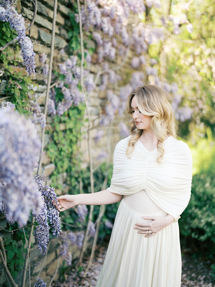 Blonde Mother In White Dress Places One Hand On Her Belly And One On Wisteria Along A Stone Wall. 