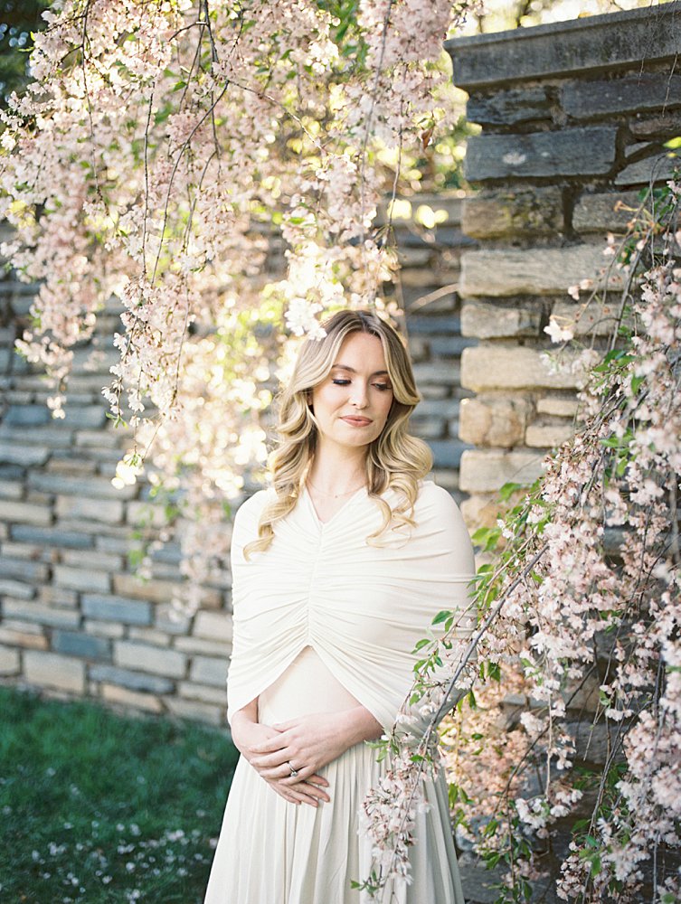A Blonde Pregnant Woman In A White Dress Stands Against A Stone Wall With Cherry Blossoms.