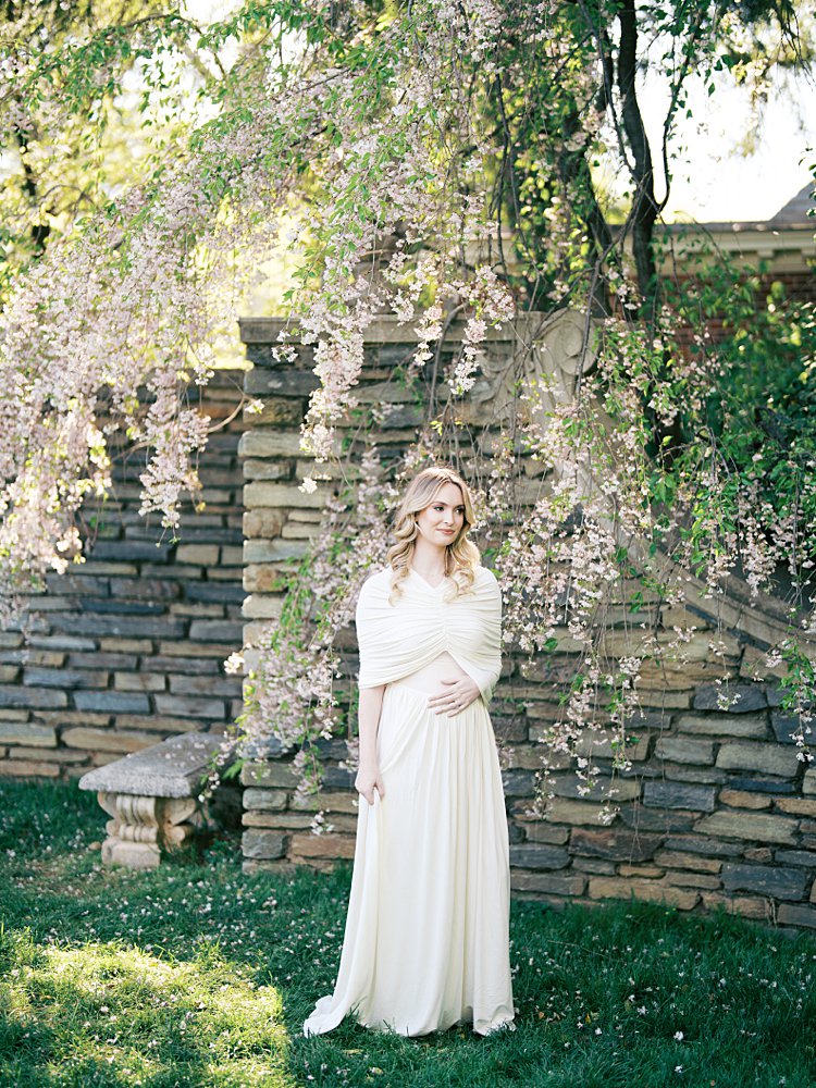 A Blonde Mother Stands Against A Stone Wall In Spring, Looking Off In The Distance, Photographed By Georgetown Photographer Marie Elizabeth Photography.