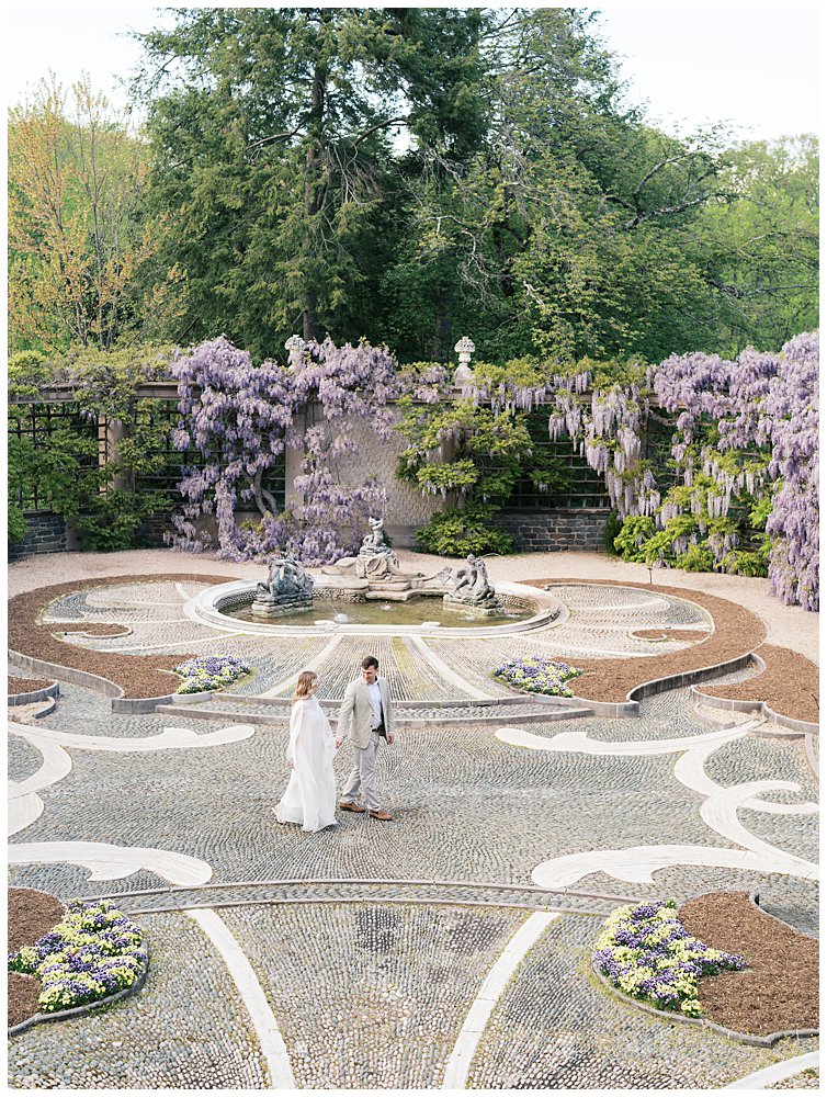A Couple Walk Across A Garden With Wisteria.