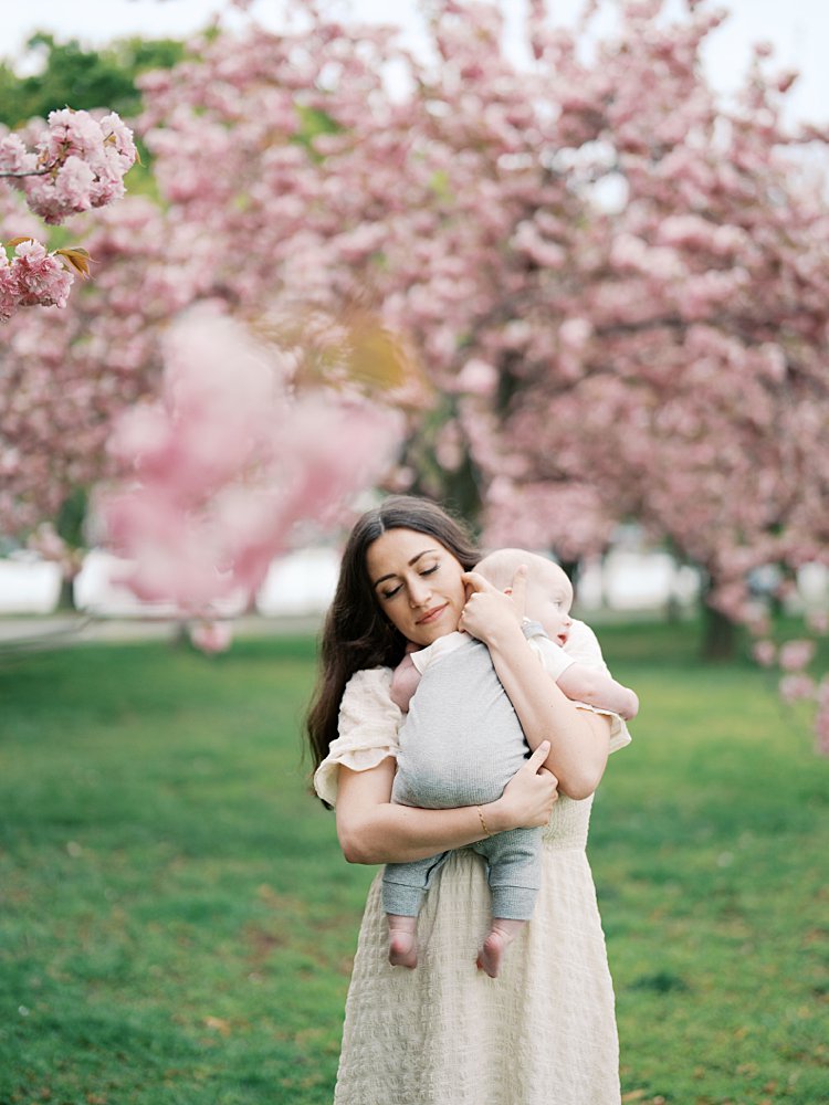 Mother With Brown Hair Holds Her Son To Her Chest During Their Family Session At The Hains Point Cherry Blossoms.
