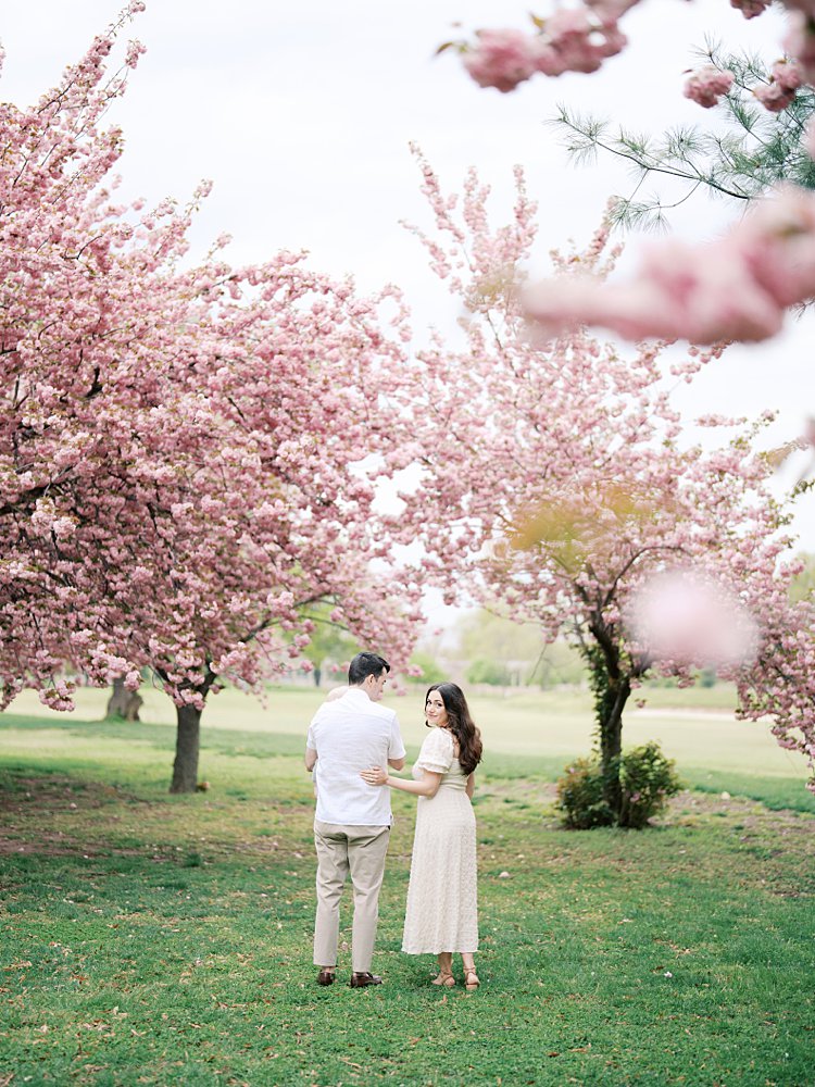 Brown Haired Woman Links Her Arm Through Her Husband's While Turning Around To Look At The Camera In A Kwanzan Cherry Tree Grove.