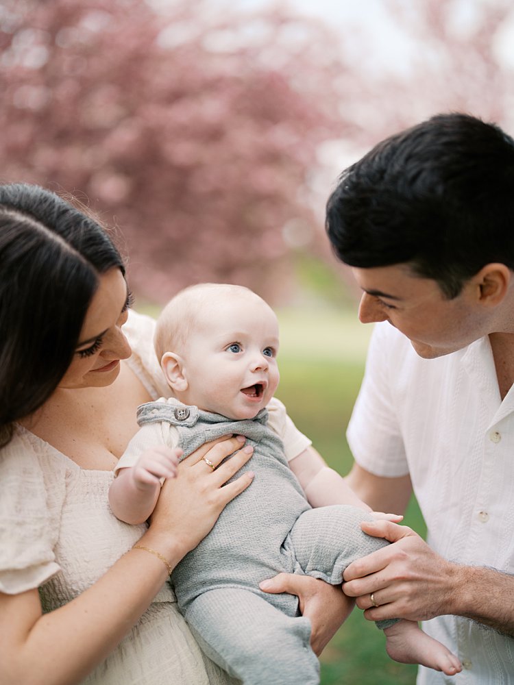 Baby Boy Smiles Up At His Father While Being Held By His Mother During Their Family Photos.