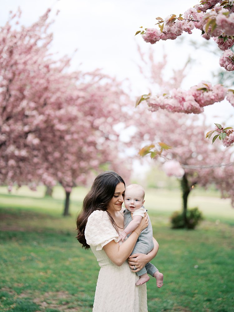 Mother With Brown Hair In White Dress Holds Infant Son Up Close To Her While Standing In The Cherry Blossoms At Hains Point In Washington Dc.