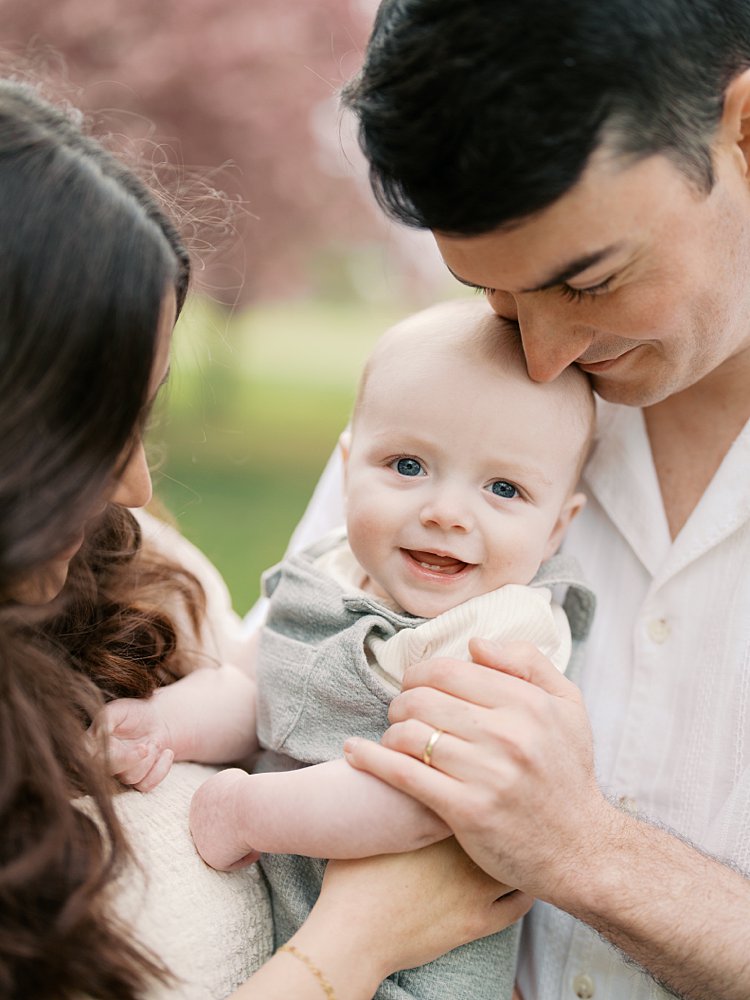 Baby Boy Smiles At The Camera While Being Held Close By His Father And Mother.