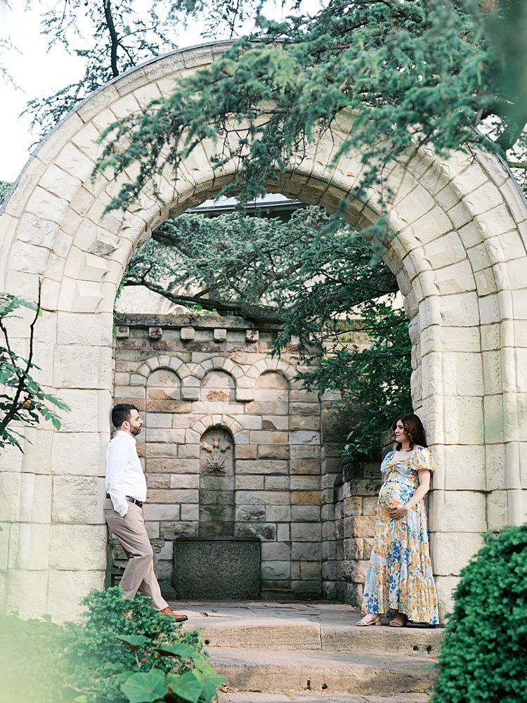 A Couple Stand Across From Each Other In A Stone Archway During Their National Cathedral Maternity Session.