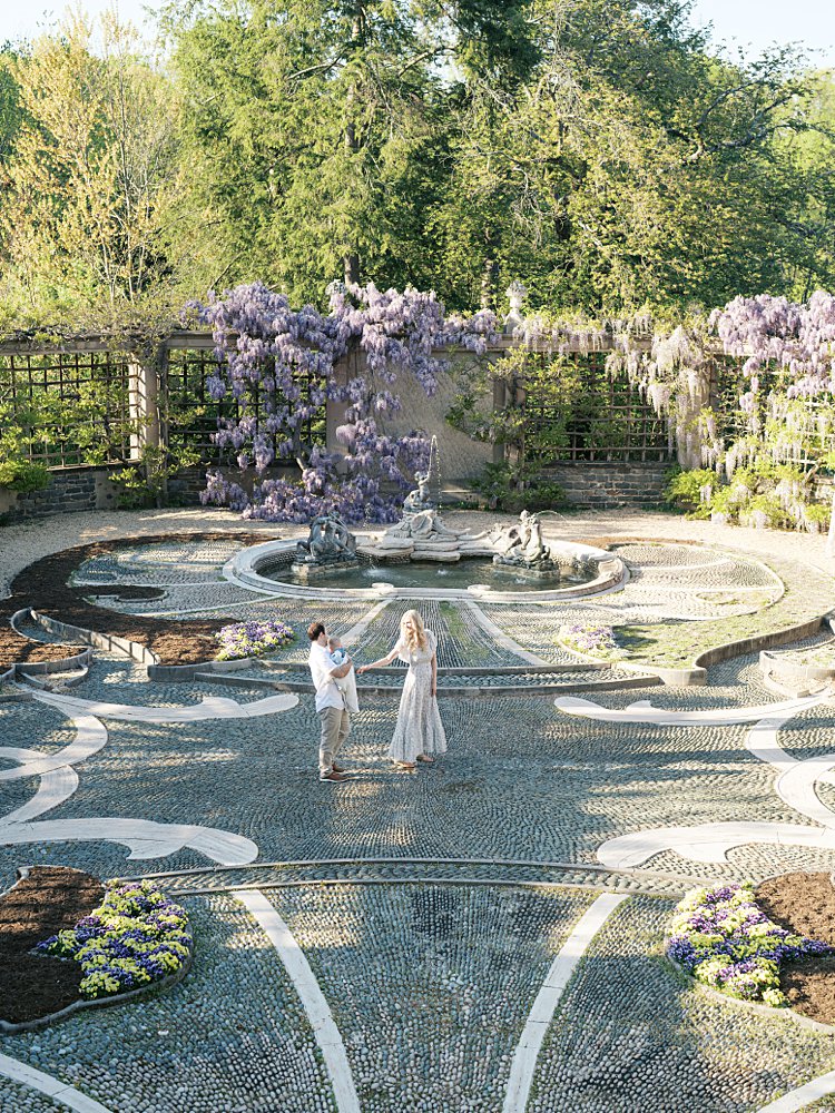 A Mother And Father Walk Across A Courtyard In Dumbarton Oaks That Is Covered In Purple Wisteria In Dc.