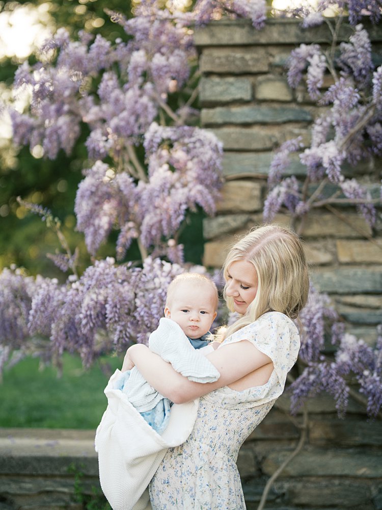 Mother Stands In Front Of Stone Wall With Hanging Purple Wisteria In Georgetown Dc, Holding Her Infant Baby Boy.