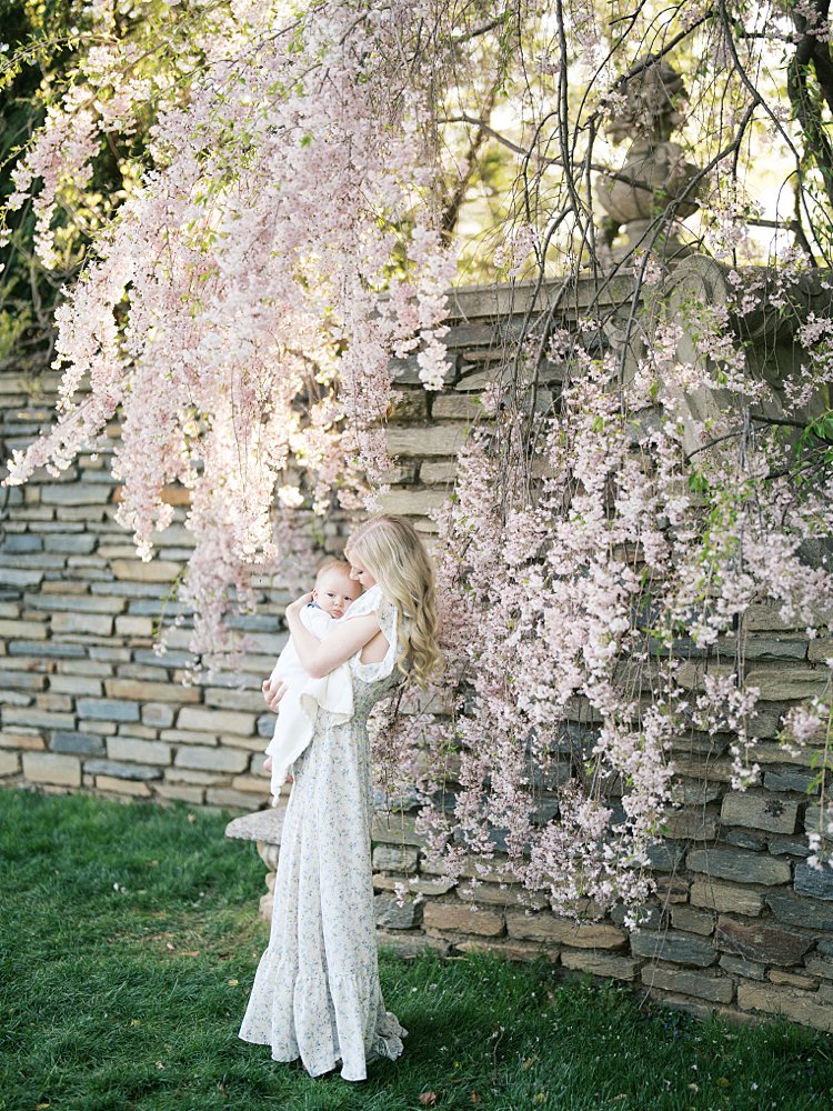 Blonde Mother Stands Near Stone Wall With Weeping Pink Cherry Tree Holding Her Baby Boy In Dumbarton Oaks In Georgetown, Dc.