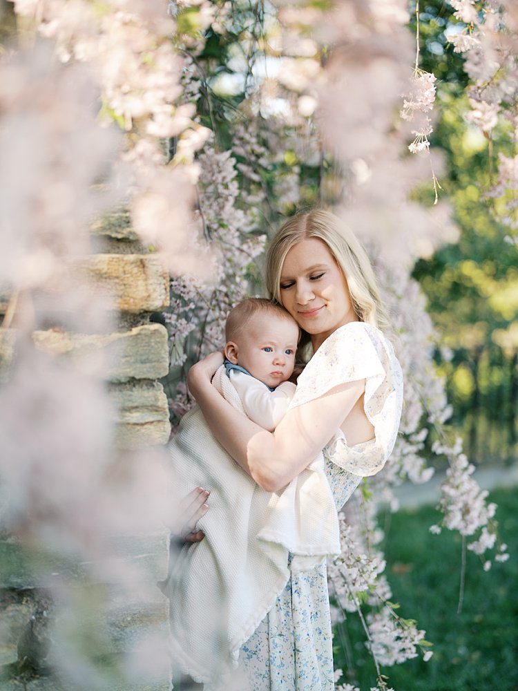 Blonde Mother Holds Her Baby Boy Close To Her While Standing In Garden With Pink Cherry Trees.