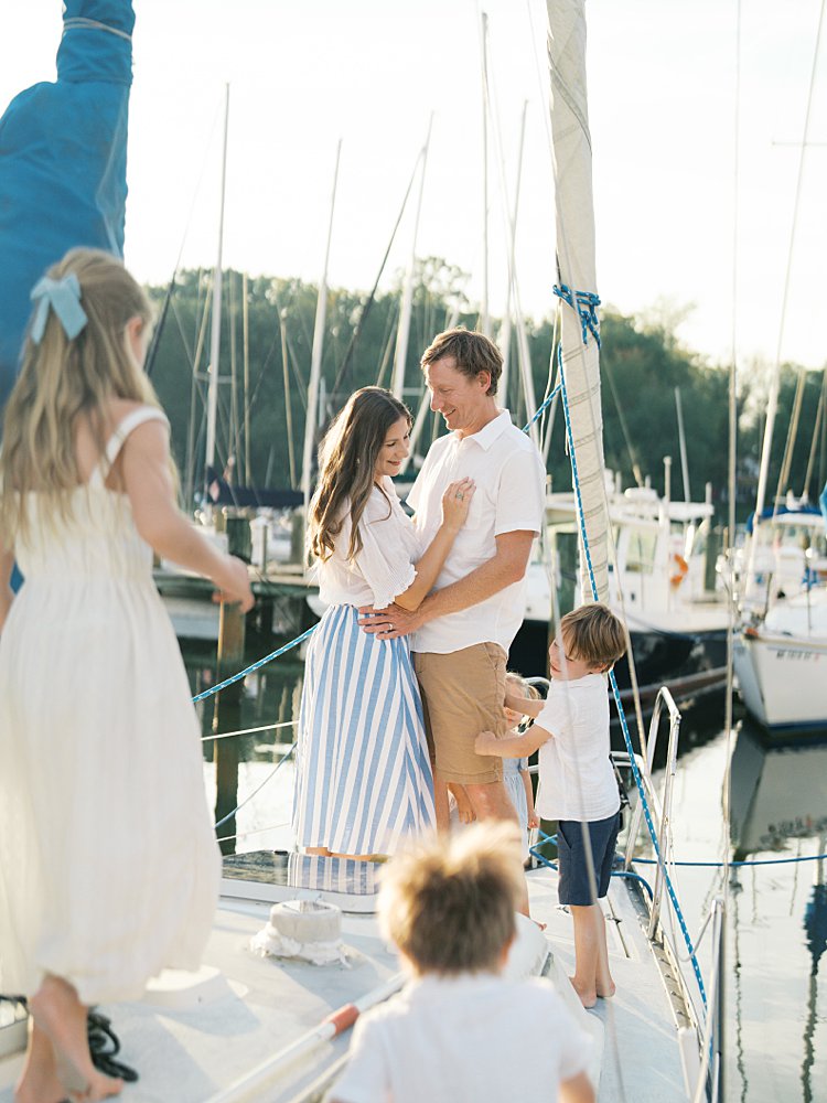 Mother and father stand arm-in-arm as their children stand along the sailboat's bow, walking towards them, photographed by Annapolis Family Photographer, Marie Elizabeth Photography.