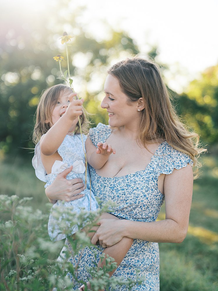 A Blonde Mother Smiles At Her Daughter As She Holds A Yellow Flower Up. 