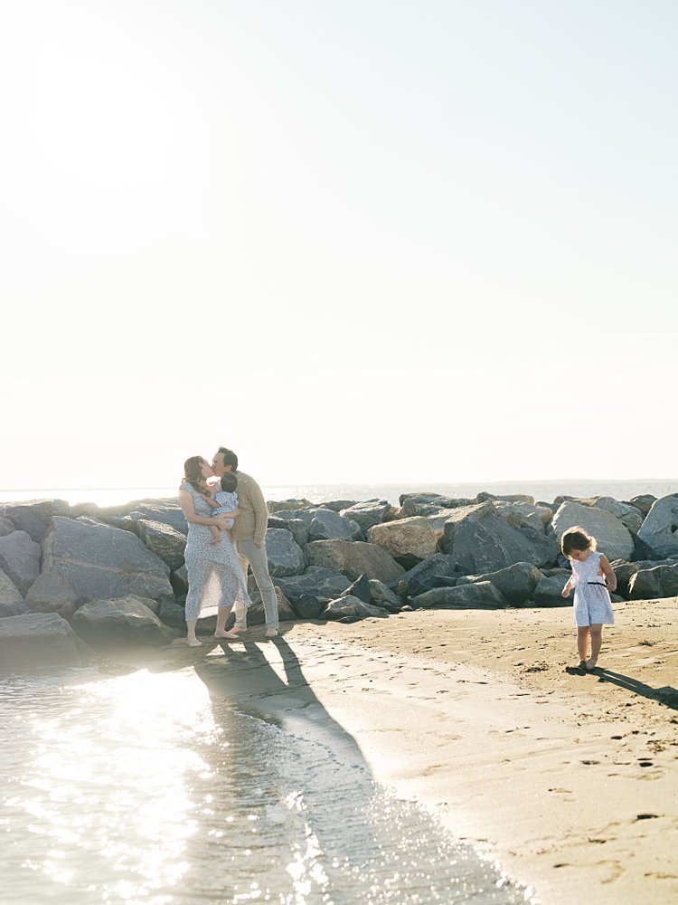 A Mother And Father Kiss As Their Two Young Children Play During Their Mayo Beach Park Photos.